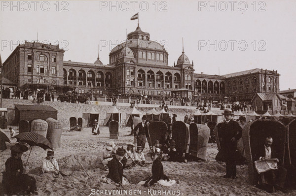 View of the Kurhaus in Scheveningen with bathers in the foreground, Scheveningen - Kurhaus., photograph, cabinet photograph, Part of a Leporello album with photographs of Scheveningen., Scheveningen, c. 1890 - c. 1910, photographer, pasteboard, ink, materials by function, paper, printing ink, collotype, photogravure, height 97 mm x width 147 mm, beach (as place of recreation), watering-place, spa, Kurhaus (Scheveningen), A wide beachfront scene shows a large ornate building with multiple arched windows, domed roof elements, and a flag atop a central cupola, sitting behind a raised promenade lined with a row of tents and a balustrade; the sandy foreground is filled with many wicker beach chairs, groups of people sitting and standing, some holding parasols, and several men in suits and hats, with one man seated reading a paper inside a chair-like shelter.