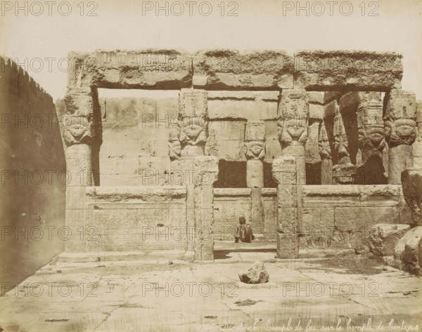 Dendérah, Temple de feu sur le temple de Tentyris, Temple complex of Dendera, photograph, Part of Travel album with 80 photos of Egypt., negative number: '125', photographer: Maison Bonfils, Dendera, 1879, North Africa, pasteboard, paper, albumen print, height 220 mm x width 279 mm, temple, shrine, Egyptian religion, A large stone facade of an ancient structure fills the frame, showing worn columns and lintels carved with decorative reliefs and symbols, some tops of columns shaped with stylized heads. The stone surfaces display weathering and textured erosion, with rectangular blocks forming the wall behind the columns. A person sits on a step within a recessed doorway in the center of the composition, wearing layered garments and a head covering. The foreground shows a paved stone courtyard with a rough rock placed near the middle and scattered debris on the ground.
