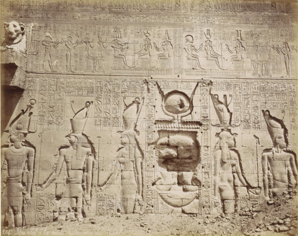 Bas-relief of the temple complex of Dendera, Bas-relief à la partie postériaire du temple Dendérah, photograph, Part of Travel album with 80 photos of Egypt., negative number: '206', photographer: Maison Bonfils, Dendera, c. 1870 - c. 1890, North Africa, pasteboard, paper, albumen print, height 214 mm x width 277 mm, piece of sculpture, reproduction of a piece of sculpture, A stone wall covered in carved bas-relief figures and decorative panels, showing seated and standing humanlike figures in profile wearing tall headdresses and holding staffs, flanked by smaller standing attendants and animal-headed figures, with rows of carved motifs and repeating symbols filling the spaces between and above the figures; large rounded carved forms and recessed circular elements appear in the central area, while a carved lion head protrudes from the upper left corner and a rough ground of scattered stones lies at the base.