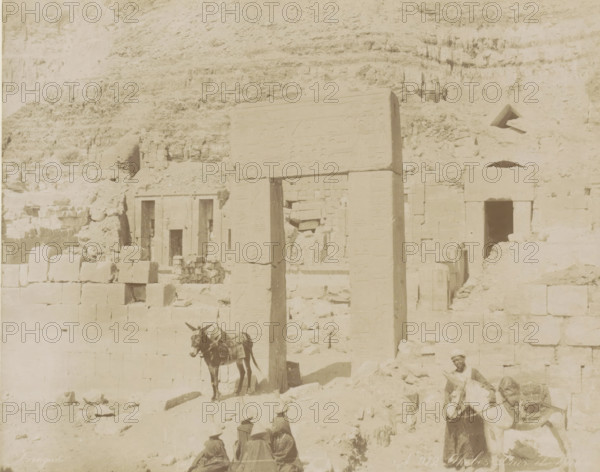 People and donkeys at the temple complex of Deir el-Bahri, Thebes Dair el Ba, photograph, Part of Travel album with 80 photos of Egypt., negative number: '912', photographer: Zangaki, C. & G., Deir el-Bahri, c. 1870 - c. 1890, North Africa, pasteboard, paper, albumen print, height 216 mm x width 275 mm, temple, shrine, Egyptian religion, A sunlit scene of weathered stone ruins set against a rocky cliff, showing a tall carved stone gateway with vertical columns and a horizontal lintel bearing shallow reliefs, adjacent stone blocks and partially intact walls with rectangular doorways and small openings, rubble and scattered stones on the uneven ground, a donkey standing near the gateway with packs on its back, a man holding another donkey near the right edge wearing light clothing and a head covering, and three seated figures wearing conical hats in the foreground.
