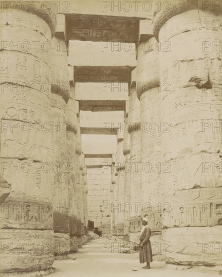 Temple of Karnak, Thebes (Luxor), Man in a colonnade with hieroglyphs (former title), photograph, Part of Travel album with 80 photos of Egypt., negative number: '1011', photographer: Zangaki, C. & G., Egypt, c. 1870 - c. 1890, North Africa, pasteboard, paper, albumen print, height 275 mm x width 220 mm, colonnade, Egyptian hieroglyphs, Luxor, A sepia-toned photograph showing a row of large, cylindrical stone columns with carved reliefs and rounded bands, forming a colonnade that recedes into the distance beneath horizontal stone lintels, with a tall stone wall visible at the far end and a single man in traditional clothing and a head covering standing barefoot near the foreground looking toward the columns.