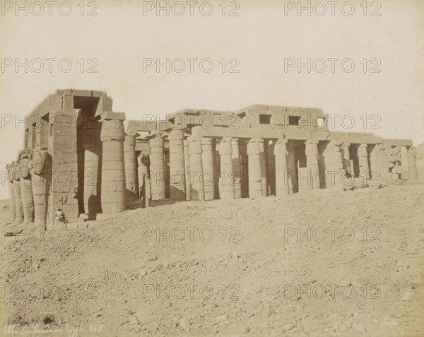 Thebes Le Ramesseum Egypt, Exterior of the Ramesseum in Thebes (Luxor), photograph, Part of Travel Album with 80 Photos of Egypt., negative number: '165', photographer: Maison Bonfils, Thebes, c. 1870 - c. 1898, North Africa, Upper Egypt, pasteboard, paper, albumen print, height 221 mm x width 280 mm, temple, shrine, Egyptian religion, A wide view of an ancient stone structure with a colonnade of tall, rounded columns supporting horizontal lintels, several carved statues and reliefs visible on the leftmost pillars, a large entrance framed by massive blocks, rubble and sandy ground in the foreground, and a lone seated figure near the base of the leftmost statues.
