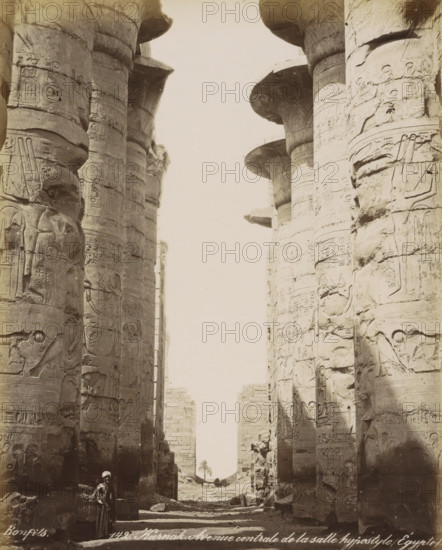 Karnak. Avenue centrale de la salle hypostyle, Egypt, Columns with hieroglyphs of the Great Hall (Hypylica Hall) in the Temple of Karnak, photograph, Part of Travel Album with 80 Photos of Egypt., negative number: '142', photographer: Maison Bonfils, Temple of Karnak, c. 1870 - c. 1898, North Africa, Luxor, pasteboard, paper, albumen print, height 279 mm x width 221 mm, temple, shrine, Egyptian religion, Egyptian hieroglyphs, A tall, narrow view between massive stone columns carved with raised figures and decorative bands, the column tops widening into rounded capitals, a worn stone pathway between the columns leading to a distant view of a low stone wall and a single palm tree, scattered stones and rubble on the ground, a person wearing a long garment and a head covering standing near the base of a column in the foreground, the scene rendered in a sepia-toned monochrome.