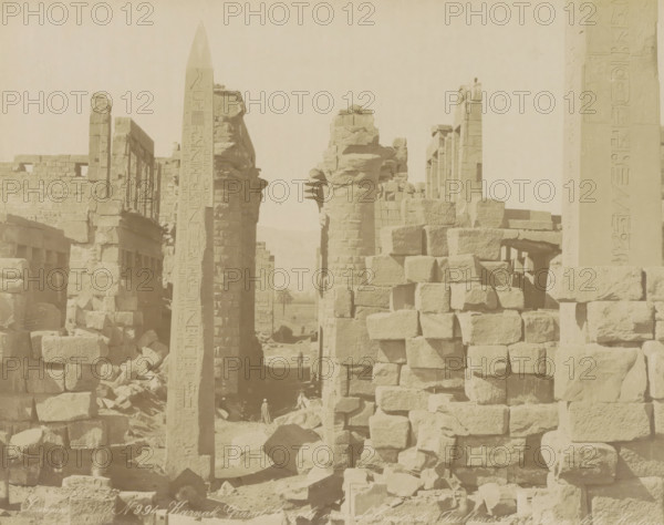 Karnak Grand temple with obélisques de Touthmes 1er et de sa fille Hatasou, Great Temple with the obelisks of Thutmose I and his daughter Hatshepsut in Karnak, photograph, Part of Travel album with 80 photos of Egypt., negative number: '994', photographer: Zangaki, C. & G., Luxor, c. 1870 - c. 1890, North Africa, pasteboard, paper, albumen print, height 216 mm x width 273 mm, obelisk, needle, temple, shrine, Egyptian religion, A wide view of an ancient stone ruin with tall carved columns and an obelisk standing among stacks of large rectangular blocks, weathered surfaces and broken masonry visible throughout, a narrow passageway between the structures leading to a distant palm tree, a few small human figures near the base of the ruins, and the scene rendered in a muted, sepia tone.