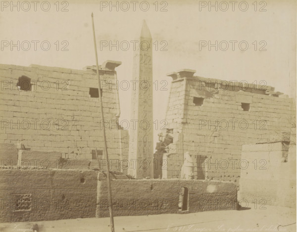 Louqsor Le pylon avec l'obelisque, Pylon with obelisk in Luxor, photograph, Part of Travel album with 80 photos of Egypt., negative number: '971', photographer: Zangaki, C. & G., Luxor, c. 1870 - c. 1890, Zangaki, photographer, North Africa, pasteboard, paper, albumen print, height 217 mm x width 276 mm, obelisk, needle, gate, entrance, A sepia-toned photograph shows a tall obelisk with carved vertical panels rising between two ancient stone walls topped with flat cornices, the walls marked by rectangular openings and surface carvings. A vertical pole leans in front of the left wall and a fragment of a statue stands near an entrance at the base of the right wall. The foreground includes a lower textured masonry wall with a small arched doorway and geometric lattice opening, and scattered stones and rubble rest along the base of the structures. The sky above is pale and empty.