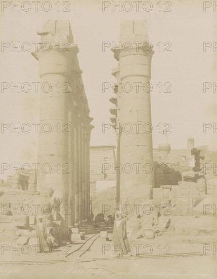 Temple of Luxor, Louksor Grande colonnade prize de profil, Colonnade with rails in Luxor (former title), photograph, Part of Travel album with 80 photos of Egypt., negative number: '976', photographer: Zangaki, C. & G., Luxor, c. 1870 - c. 1890, North Africa, pasteboard, paper, albumen print, height 276 mm x width 218 mm, colonnade, railway, train, Two tall stone columns with carved capitals stand in the foreground, forming a passageway between them that leads toward a stone wall and ruins beyond. The columns show horizontal joint lines and sculpted projecting elements along their shafts. Flanking the passage are scattered stone blocks and rubble, some stacked into low walls and broken structures. A narrow set of rails runs along the ground through the scene, partially buried among stones. Several figures in long robes and head coverings stand and sit near the ruins, their faces and postures visible but not detailed. In the distant background are more stone structures and a tower with ...