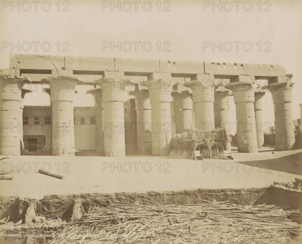 Grande colonnade de Louqsor (Égypte), Temple of Luxor, Great colonnade in Luxor (former title), photograph, Part of Travel Album with 80 Photos of Egypt., negative number: '127', photographer: Maison Bonfils, Luxor, c. 1870 - c. 1898, North Africa, pasteboard, paper, albumen print, height 224 mm x width 280 mm, colonnade, A row of large stone columns with flared capitals supports a heavy horizontal lintel, forming an open colonnade in front of a stone building with rectangular window openings; a small person sits at the base of one column; bundles of reeds or thatch are stacked against part of the colonnade near a simple woven shelter; scattered logs and sticks lie on the sandy ground in the foreground, where an exposed trench edge reveals masonry and piled wooden poles below.