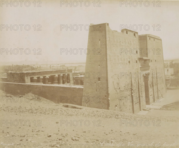 Edfou Vue general e du temple, View of the Temple of Horus in Edfu, photograph, Part of Travel album with 80 photos of Egypt., negative number: '1025', photographer: Zangaki, C. & G., Temple of Horus (Edfu), c. 1870 - c. 1890, North Africa, pasteboard, paper, albumen print, height 219 mm x width 268 mm, temple, shrine, Egyptian religion, A large stone structure with tall, sloping walls covered in carved figures occupies the right side of the image, showing doorways and reliefs and a small projecting balcony, while to the left a row of columns and a low surrounding wall sit behind a sandy foreground scattered with small stones and depressions, and in the distant background there is a flat landscape with low vegetation and a few taller trees under a pale sky.