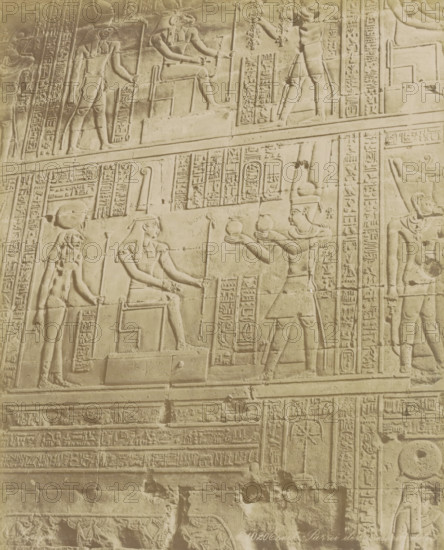 Esneh Parroi der, de te, Wall with hieroglyphs and reliefs in Esna, photograph, Part of Travel album with 80 photos of Egypt., negative number: '1020', photographer: Zangaki, C. & G., Esna, c. 1870 - c. 1890, North Africa, pasteboard, paper, albumen print, height 272 mm x width 220 mm, sculpture (relief, sculpture), Egyptian hieroglyphs, A carved stone wall panel showing raised relief figures and decorative bands, with multiple humanlike figures in profile wearing headdresses and holding staffs or offering vessels, seated and standing poses visible, framed by vertical and horizontal panels filled with repeated pictorial symbols, the surface showing signs of wear and faint discoloration, and rows of ornamental carved borders separating different registers of the scene.