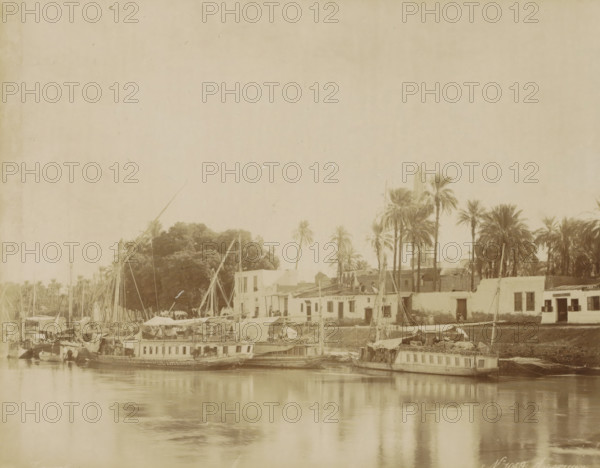 Assouan, Ships (dahabies) at a quay in Aswan, photograph, Part of Travel album with 80 photos of Egypt., negative number: '1049', photographer: Zangaki, C. & G., Aswan, c. 1870 - c. 1890, North Africa, pasteboard, paper, albumen print, height 219 mm x width 276 mm, sailing-ship, sailing-boat, A riverside scene shows several wooden boats moored along a calm waterway with reflections on the surface; behind the boats are low buildings with flat roofs and small windows, some with open shutters and a few rooftop structures; tall slender palm trees and a cluster of larger trees rise behind the buildings, and a taller tower-like structure is visible among the trees; rigging, masts, and sails extend upward from the boats, and various awnings, canopies, and cargo are visible on deck; the overall image has a sepia tone and an aged paper texture.