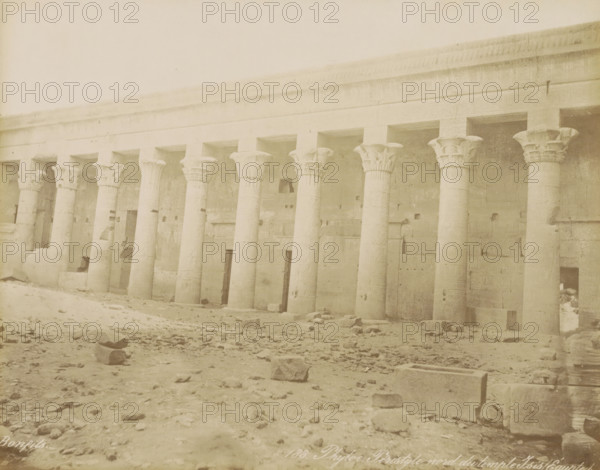 Phyloe. Peristyle nord du temple Isis (Egypt), Northern colonnade (peristilium) of the Temple of Isis on the island of Philae, photograph, Part of Travel album with 80 photos of Egypt., negative number: '183', photographer: Maison Bonfils, Philae, c. 1870 - c. 1898, North Africa, pasteboard, paper, albumen print, height 221 mm x width 280 mm, colonnade, temple, shrine, Egyptian religion, Temple of Isis (Philae), A wide exterior view of a long stone colonnade with tall carved columns topped by ornate capitals, each column set into a recessed bay beneath a continuous entablature, a rough stone wall behind the columns showing shallow niches and rectangular openings, a sandy and rubble-strewn foreground with scattered blocks and debris, and a pale sky above.