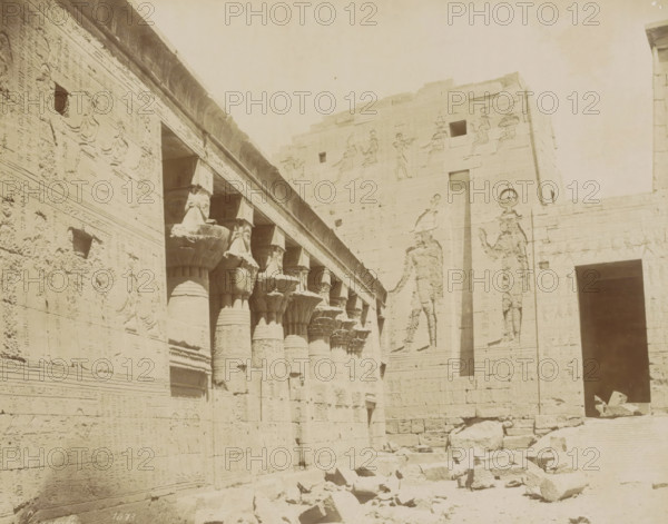 Colonnade of the Temple of Isis on the island of Philae, photograph, Part of Travel album with 80 photos of Egypt., negative number: '1073', photographer: Zangaki, C. & G., Philae, c. 1870 - c. 1890, North Africa, pasteboard, paper, albumen print, height 215 mm x width 274 mm, colonnade, temple, shrine, Egyptian religion, Temple of Isis (Philae), A sunlit stone facade of an ancient structure with rows of carved columns topped by floral and bud-shaped capitals, a colonnade running diagonally into the image, and detailed bas-relief figures and motifs carved into the walls; fragments of stone and rubble lie scattered on the ground in front of the building, and a large doorway or opening is visible on the right with shadowed interior space.