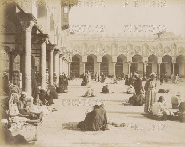 Students in the courtyard of the Al-Azhar Mosque in Cairo, Courtyard and colonnade of the Al-Azhar Mosque, with students, photograph, Part of a travel album with 80 photos of Egypt., negative number: '1371', photographer: Maison Bonfils, Cairo (city), c. 1870 - c. 1898, Ottoman Empire, North Africa, pasteboard, paper, albumen print, height 224 mm x width 280 mm, temple, shrine, Islam, Mohammedanism, A wide sunlit courtyard bordered by a building with a row of tall arches and decorative round medallions above each arch, and a colonnaded portico with columns and carved capitals along one side; many people wearing long robes and head coverings sit, stand, walk, or recline across the paved ground, some grouped near the columns and others scattered toward the center and far side; shadows from the columns fall across the courtyard; a few hanging lamps and ornate bracketed fixtures are attached to the portico; architectural details include carved patterns along the upper wall and a projecting...