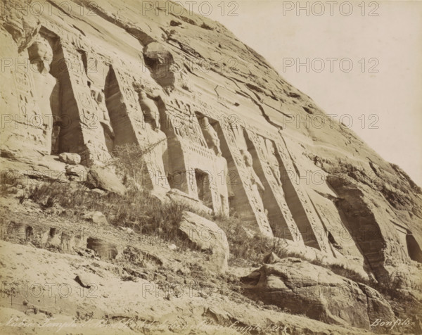 Nubie. Temple d' Abou Simbel, dédie a Athor (Égypt), Temple of Abu Simbel, photograph, Part of Travel album with 80 photos of Egypt., negative number: '201', photographer: Maison Bonfils, Abu Simbel, c. 1870 - c. 1898, Maison Bonfils, photographer, North Africa, Upper Egypt, pasteboard, paper, albumen print, height 221 mm x width 279 mm, sculpture (relief, sculpture), temple, shrine, Egyptian religion, A steep rock face with carved vertical recesses and sculpted humanlike figures integrated into the stone, fluted columns and rectangular doorways set into the cliff, patterned reliefs decorating the carved surfaces, scattered boulders and ledges at the base, sparse dry vegetation growing among the rocks, and a pale sky above.