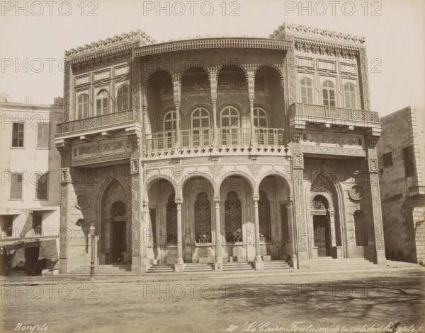 Le Caire, Fontaine de la Validé (Egypt), Building with well in Cairo, photograph, Part of Travel Album with 80 Photos of Egypt., negative number: '80', photographer: Maison Bonfils, Cairo (city), c. 1870 - c. 1898, Ottoman Empire, North Africa, pasteboard, paper, albumen print, height 220 mm x width 281 mm, utilitarian, public pump or water fountain, A frontal view of a large ornate building facade with an upper level featuring rows of arched windows and decorative railings, and a lower level with a series of tall arches supported by slender columns leading to intricately patterned window grilles and doorways; the facade displays carved and geometric ornamentation around arches and windows, latticework balconies, and decorative friezes along the top edge; steps lead up to the entrance area and a street lamp stands near one side; adjacent buildings with shuttered windows flank the ornate structure and a bare ground surface occupies the foreground.