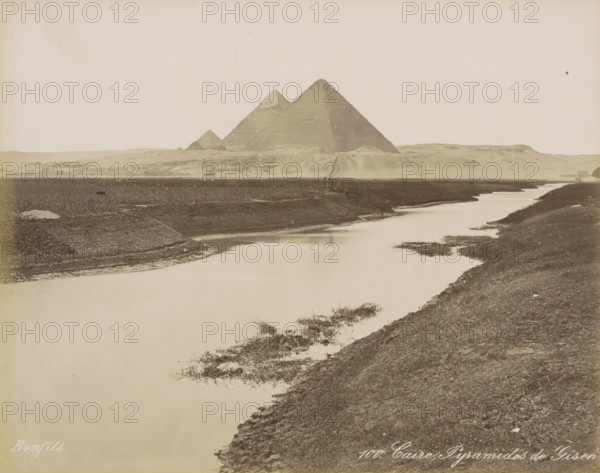 Caire, Pyramids of Giza, View of the Pyramids of Giza near Cairo, photograph, Part of Travel Album with 80 Photos of Egypt., negative number: '100', photographer: Maison Bonfils, Giza, c. 1870 - c. 1898, North Africa, pasteboard, paper, albumen print, height 220 mm x width 282 mm, pyramid (historical grave form), Ottoman Empire, Cairo (city), A wide sepia-toned landscape showing a calm winding river in the foreground with grassy and muddy banks, a low flat plain beyond the river, and three large pyramidal stone structures on a distant rise against a pale sky, with sparse vegetation and a small boat or wooden structure near the far bank.
