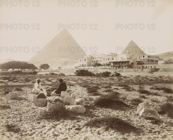 Hotel Mena - House and pyramids, View of the Pyramids of Giza with Hotel 'Mena House' in the foreground, photograph, Part of Travel Album with 80 Photos of Egypt., negative number: '1434', photographer: Maison Bonfils, Giza, c. 1886 - c. 1898, North Africa, pasteboard, paper, albumen print, height 221 mm x width 272 mm, hotel, hostelry, inn, pyramid (historical grave form), A wide sandy landscape with scattered low shrubs and rocks in the foreground shows two seated figures on a cluster of stones, one wearing a light robe and head covering and the other in darker clothing and a brimmed hat, facing each other. Behind them a built cluster of multi level stone and stucco buildings with flat roofs and a small domed structure runs along a raised embankment, with trees visible to the side. Two large triangular stone pyramids rise in the background behind the buildings, occupying much of the sky area. The overall scene is sepia toned with textured sand and sparse vegetation across the foregro...