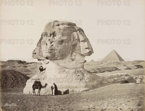 Sphinx and pyramids of Giza, Caire, Sphynx et Pyramide, photograph, Part of Travel album with 80 photos of Egypt., negative number: '111', photographer: Maison Bonfils, Gizeh, c. 1870 - c. 1898, North Africa, Giza, pasteboard, paper, albumen print, height 220 mm x width 282 mm, pyramid (historical grave form), piece of sculpture, reproduction of a piece of sculpture (sculpture), A large weathered stone sculpture of a human head and neck dominates the foreground, showing layered textures and erosion lines on its surface. A person sits on the stone near the sculpture’s base, and another person stands in front of the base beside a camel, which has a saddle and some coverings. A small figure is positioned on a ledge on the sculpture’s neck. Low rocky hills and sandy ground surround the sculpture, and a pyramid with a smooth sloped face rises in the distant background against a pale sky.