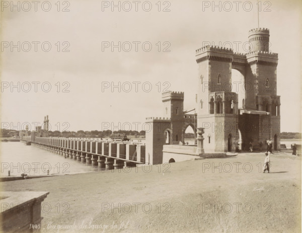 View of a dam on the Nile near Cairo, General view of the Nile dam, photograph, Part of a travel album with 80 photos of Egypt., check stamp: '1445', photographer: Maison Bonfils, Egypt, c. 1870 - c. 1898, North Africa, pasteboard, paper, albumen print, height 221 mm x width 281 mm, dam, Ottoman Empire, Cairo (city), A wide riverside scene shows a long bridge extending into the distance with evenly spaced vertical supports rising from the water, leading to a series of brick towers with crenellated tops and arched openings; a larger ornate brick gateway with rounded turrets and decorative arches stands near the foreground, showing carved stonework and columned openings, while people in period clothing walk and stand near the entrance and on the embankment; a grassy slope occupies the right foreground and the calm river fills the left foreground, with a distant tree-lined shore visible under a pale sky.