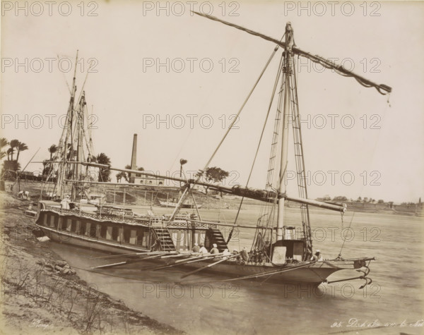 Dahabie on the Nile, Passenger boat (dahabie) on the Nile, photograph, Part of Travel Album with 80 Photos of Egypt., negative number: '95', photographer: Maison Bonfils, Egypt, c. 1870 - c. 1898, North Africa, pasteboard, paper, albumen print, height 222 mm x width 278 mm, sailing-ship, sailing-boat, Nile, A large boat with tall masts and long spars is docked along a riverbank, showing a wooden hull with a row of cabin windows and an open deck with railings and steps leading down; several ropes, rigging lines, and ladders run up the masts, and a group of people wearing light clothing and head coverings sit or stand near the stern and along the deck; the river water is calm and stretches across the image with another boat partly visible in the background; palm trees and a building with a tall chimney stand on the far shore, and the sky above is clear and pale.