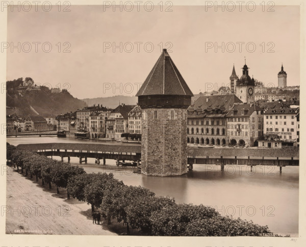 View of the water tower in Lucerne, album page, photomechanical print, This print is part of an album., inscription: 'Edit. Schroeder & Cie., Zürich, 76. Luzern, Wasserthurm', annotation: '1897', publisher: Schroeder & Cie., Zürich (city), 1897, paper, collotype, photoprint, height 265 mm, width 350 mm, city-view in general, 'veduta', water tower, Luzern, A riverside scene in sepia tones showing a large stone tower with a steep, shingled pyramidal roof rising from the water beside a covered wooden bridge that spans the river; rows of multi-story buildings with shuttered windows and arched arcades line the far bank behind the tower, and a clock tower and other steepled towers rise among the rooftops; a tree-lined promenade runs along the near bank with a few people standing beneath the trees, and hills with scattered buildings are visible in the background.