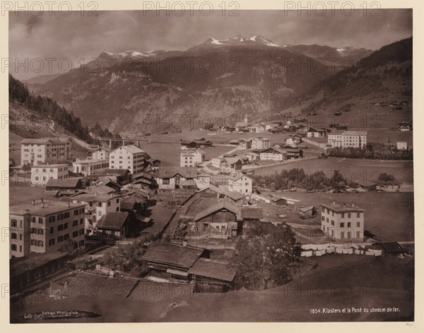 View of Klosters with railway bridge, album page, photomechanical print, This print is part of an album., annotation: '1897', inscription: 'Edition Photoglob, 1654. Kloster et le Pont du chemin de Fer', publisher: Photoglob & Co., 1897, paper, collotype, photogravure, height 265 mm, width 350 mm, city-view in general, 'veduta', railway, tramway, rack railway, Graubünden (canton), A sepia-toned panoramic view shows a valley village with clustered buildings of varying sizes and styles, including multi-story rectangular houses and smaller wooden structures with sloped roofs. In the foreground there are rows of rooftops, fenced yards, and some laundry hung out to dry. A mix of open grassy fields and cultivated plots lie between the buildings, with narrow roads or paths winding through the settlement. Further back a larger open area is bordered by more houses and a prominent towered building. Beyond the village the terrain rises into forested hills and steep mountains with lighter patches n...