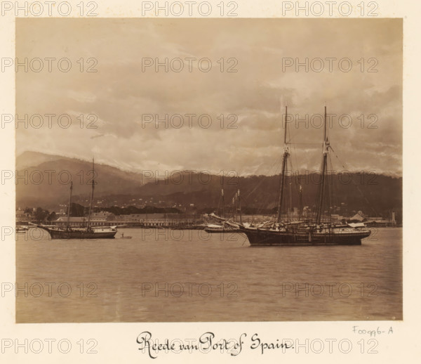 View of the harbor of Port of Spain, album page, photograph, This photo is part of an album., inscription: 'Reede van Port of Spain', Trinidad (Trinidad and Tobago), 1880 - 1900, photographer, paper, albumen print, height 347 mm x width 297 mm, city-view in general, 'veduta', harbor (view, vehicle, ship, etc.), A sepia-toned photograph showing a calm harbor scene with sailing ships moored on the water, tall masts and rigging rising against a cloudy sky, a long waterfront of low buildings along the shore, hills in the background partially covered by clouds, and reflections of the vessels and sky visible on the water surface.