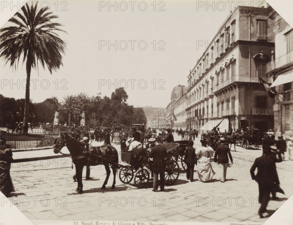 Street Naples, Napoli Riviera di Chiaja e Villa Nazionale, Street in Naples with horse-drawn carriages and passers-by, photograph, Part of Album with 191 photos of a trip to and through South Africa., negative number: '13', Naples, publisher: Edizione Esposito, c. 1880 - c. 1920, photographer, paper, albumen print, albumendruk, height 198 mm x width 258 mm, four-wheeled, animal-drawn vehicle, eg: cab, carriage, coach, street (city(-scape) with figures, staffage), A wide street scene with a large palm tree and fenced garden area on the left and a long row of multi-story buildings with balconies and awnings on the right, crowded with people, horse-drawn carriages, and horses filling the foreground and midground; pedestrians in period clothing walk and stand along the street, some near the carriages, while lamp posts and statues are visible within the garden area and trees rise behind the fencing; the paved street extends into the distance where more buildings and small distant figures ar...