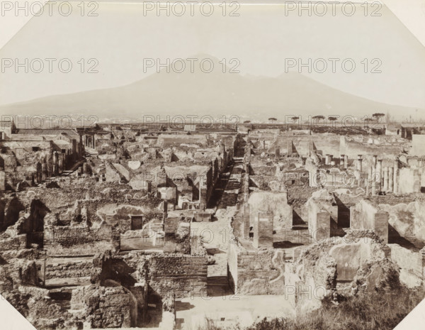 View of the ruins of Pompeii at the foot of Mount Vesuvius, Pompei: panorama e Vesuvio, City of Pompei, photograph, Part of Album with 191 photos of a trip to and through South Africa., negative number: '65', Pompeii, publisher: Edizione Esposito, c. 1880 - c. 1920, photographer, paper, albumen print, height 200 mm x width 259 mm, ruin of a dwelling, house, castle, Mount Vesuvius, A wide sepia-toned view of extensive ancient ruins fills the foreground with fragmented stone walls, columns, and archways forming irregular room outlines and narrow paved streets running toward the middle distance, where a low wall and scattered trees mark a boundary; beyond that, a large mountain rises across the horizon under a pale sky, its slopes gently sloping to smaller peaks at the sides.