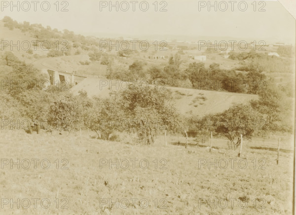 Pretoria, Landscape with railway line near Pretoria, photograph, Part of Album with 191 photos of a trip to and through South Africa., c. 1890 - c. 1920, photographer, paper, daylight gelatin silver print, height 213 mm x width 293 mm, railway, train, A wide pale landscape with rolling hills and scattered trees dominates the scene, with a curving dirt track leading to a low stone bridge spanning a shallow ravine; a few small buildings and structures appear in the distant midground beyond the bridge, and clusters of trees and shrubs fill the slopes and valley below; in the foreground a lone seated man in a hat sits near a bush on grassy terrain dotted with small rocks, and a simple wooden fence with posts runs along the lower right side.