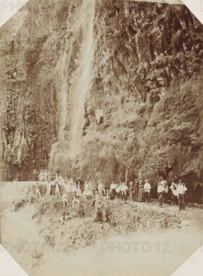 Madeira, Tourist group at a waterfall on Madeira, photograph, Part of Album with 191 photos of a trip to and through South Africa., inscription: 'Rabacal ...', c. 1885 - c. 1910, photographer, paper, albumen print, height 236 mm x width 176 mm, waterfall, A sepia-toned photograph showing a steep rocky cliff covered in grasses and vegetation with a narrow waterfall cascading down its face, and a group of people gathered on a stone path and ledges at the base of the cliff, some standing and some seated, wearing period clothing and hats, with walking sticks and a few small bags, set against the textured rock and plant-covered slope.