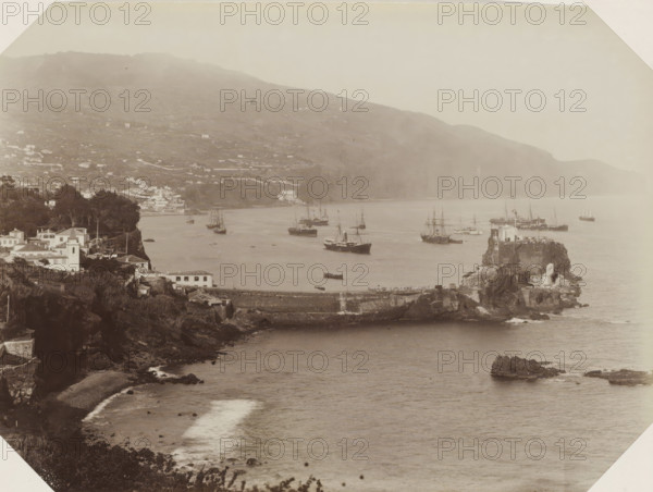 View of the coast and a harbor pier of Madeira, photograph, Part of Album with 191 photos of a trip to and through South Africa., name: '... and Look-Book Madeira', Madeira, c. 1885 - c. 1910, photographer, paper, albumen print, height 171 mm x width 232 mm, pier, quay, wharf, A wide coastal scene showing a bay with calm water and a rocky shoreline, a stone breakwater extending into the water, a rocky promontory with a small building on top, several sailing ships and steamers anchored or drifting in the bay, a cluster of white buildings and houses along a tree-lined cliff, terraced hills rising in the background, scattered vegetation and rocky outcrops along the foreground shore, and distant misty hills along the horizon.