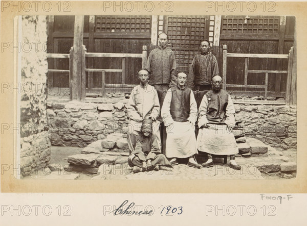 Chinese men and a boy in front of a house in Tibet, photograph, Part of Photo album with 24 photos of army telegrapher DT Dalton's journey through Tibet., title: 'Chinese 1903', photographer: DT Dalton, Tibet, 1903, DT Dalton, photographer, paper, daylight gelatin silver print, height 80 mm x width 137 mm, shoes, sandals, head-gear, child, hairdress, front steps, clothing for the upper part of the body, clothing for the lower part of the body, A sepia photograph shows a group of people posed on uneven stone steps in front of a wooden building with a lattice door and railings; two adults stand on the top step behind three seated adults on a lower step, and a child sits cross-legged at the front smiling; the adults wear long traditional garments with layered tops and wide aprons or robes, and some wear round caps while one holds a small rectangular object; the stone wall and steps have rough, irregular stones and the wooden facade behind them has vertical posts and horizontal beams with ...