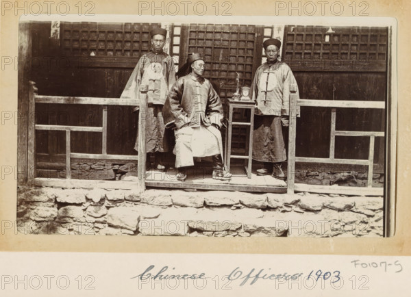 Chinese officers in front of a house in Tibet, photograph, Part of Photo album with 24 photos of army telegrapher DT Dalton's journey through Tibet., title: 'Chinese Officers 1903', photographer: DT Dalton, Tibet, 1903, DT Dalton, photographer, paper, daylight gelatin silver print, height 80 mm x width 137 mm, shoes, sandals, official, civil servant, functional (in general), head-gear, clock, clothing for the upper part of the body, clothing for the lower part of the body, A sepia-toned photograph showing three men wearing traditional long robes and round hats posed on a raised wooden platform in front of a building with wooden lattice windows, a simple wooden railing in front of them, a small table holding a few objects beside the seated man, stonework forming the base of the platform, and the men displaying neutral facial expressions.
