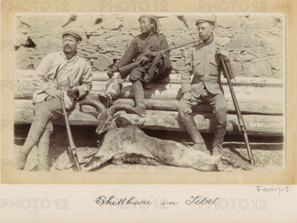 Shikhari (hunters), two Englishmen among whom possibly sergeant Dalton and hunting booty in Tibet, photograph, Part of Photo album with 24 photos of army telegraphist DT Dalton's journey through Tibet., title: 'Shikhari in Tibet', photographer: DT Dalton, Tibet, 1903 - 1906, DT Dalton, photographer, paper, daylight gelatin silver print, height 80 mm x width 137 mm, Person, spoils of the hunt, game, venison, firearms: rifle, group-portrait of hunters, clothes for special purposes, A sepia photograph of three seated people on a rough log bench in front of a stone wall, the person on the left wearing a light-colored jacket and cap with a mustache and holding a long rifle upright, the person in the center wearing darker, tattered clothing and holding a smaller firearm while resting a carcass on their lap, and the person on the right wearing a uniform jacket and cap with a rifle balanced on their shoulder; an animal with curved horns lies on the ground in the foreground, partially covering ...
