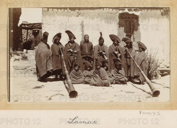 Group of Tibetan lamas with wind instruments (dunchen), Tibet, photograph, Part of Photo album with 24 photos of the journey of army telegraphist DT Dalton through Tibet., title: 'Lamas', photographer: DT Dalton, 1903 - 1906, paper, daylight gelatin silver print, height 80 mm x width 137 mm, head-gear, wind instruments, clothes covering the entire body, priest, non-Christian religions, A group of people arranged in two rows in front of a worn plaster wall, most wearing long cloaks and distinctive tall hats with fur or pointed tops, several seated on the ground wrapped in cloaks, some standing behind them holding long, ornate horns or ceremonial instruments that extend toward the camera, a narrow doorway or window with a decorative frame visible on the right side of the wall, rough stone paving underfoot and an opening to a darker stone structure at the left edge of the scene.