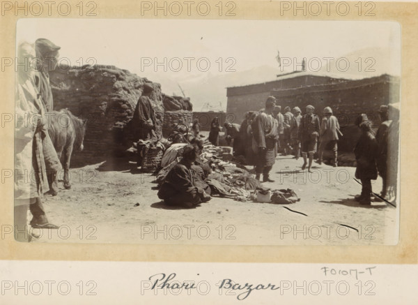 Market in Phari town, Tibet, photograph, Part of Photo album with 24 photos of the journey of army telegraphist DT Dalton through Tibet., title: 'Phari Bazar', photographer: DT Dalton, Phari, 1903 - 1906, paper, daylight gelatin silver print, height 80 mm x width 137 mm, market, buying and selling, A sepia photograph of an outdoor market scene with many people wearing layered, loose clothing and hats, some standing and some sitting on the ground among piles of textiles and baskets, a few figures clustered in conversation near a low wall, a large rounded haylike structure and a rectangular building in the background, mountains faintly visible in the distance, a small animal standing near the left edge, and fragments of a light border surrounding the image.