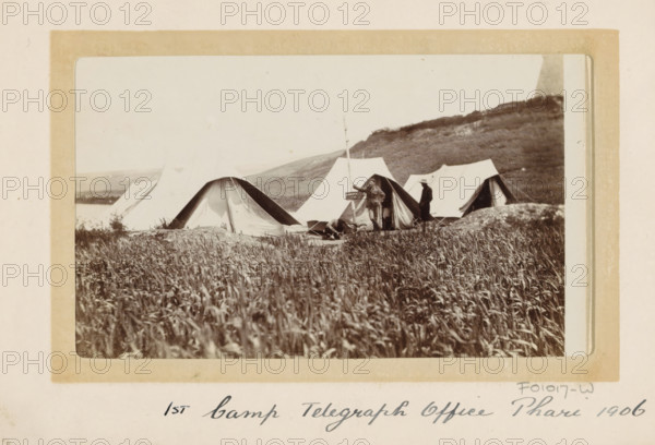 Tent camp of the first British telegraph post in Phari, Tibet, photograph, Part of Photo album with 24 photos of the journey of army telegraphist DT Dalton through Tibet., title: '1st Camp Telegraph Office Phari 1906', photographer: DT Dalton, Phari, 1906, paper, daylight gelatin silver print, height 80 mm x height 137 mm, (military) camp with tents, telegraph pole, telephone pole, (morse) telegraph, A sepia photograph of a row of canvas tents pitched on a grassy plain with taller grass in the foreground and a low hill in the background. Two men stand near one tent, one holding a long pole and the other wearing a hat, and another person lies or sits on the ground nearby. The tents vary in shape with rounded and peaked entrances, and some flaps are open revealing darker interiors. The image is mounted on a light colored card with a rectangular border around the photograph.
