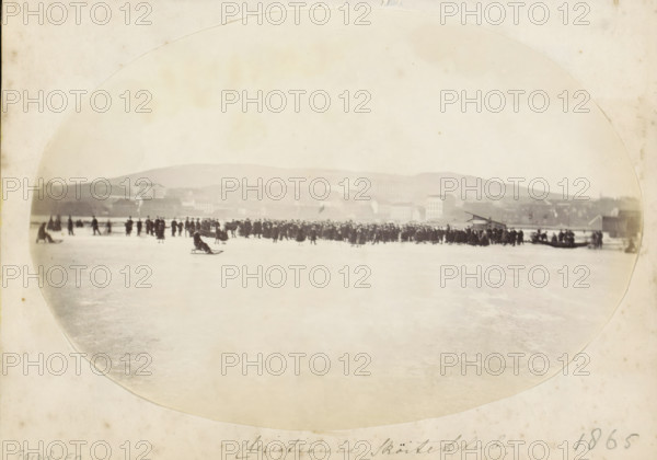 Skaters in Christiania, Christiania Sköiteklub, photograph, Part of an English family album with photos of people, travel, cricket and artworks., date: '1865', 1865, photographer, paper, albumen print, height 138 mm x width 201 mm, skates (winter sports), prospect of village, silhouette of village, Christiania, A wide, faded black-and-white scene shows a frozen expanse with many small dark figures gathered across the middle ground, some standing and some clustered in groups, while a few individuals sit on sled-like objects on the ice; buildings and low hills form a distant hazy background under a pale sky, and the image is framed by an oval vignette and surrounded by a light margin.