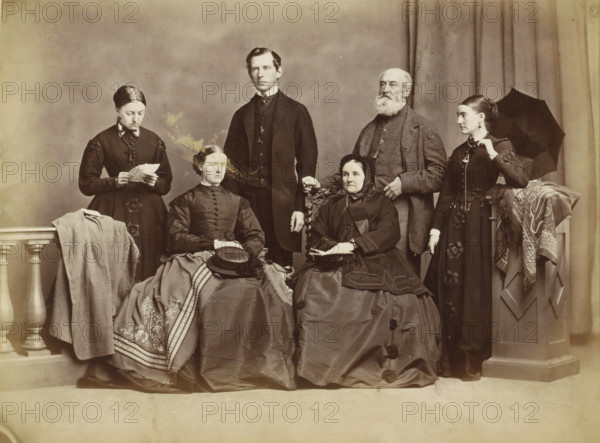 Group portrait of a family in a photo studio, possibly in Norway, photograph, Part of an English family album with photos of people, travels, cricket and artworks., inscription: '...famil...', Norway, c. 1860 - c. 1870, photographer, paper, albumen print, albumendruk, height 156 mm x width 205 mm, historical persons, family group, especially parents with their child(ren), head-gear, beard, dress, gown, skirt, clothing for the upper part of the body, styles of hairdress - AA -, women, umbrella, necklace, studio requisites, clothing for the lower part of the body, A sepia-toned portrait of six people arranged in two rows against a plain backdrop and draped curtain, with three seated women in full skirts and three standing adults behind them; the seated woman on the left wears a dark dress with decorative trim and a patterned shawl draped over her lap with a hat placed on top, the seated woman in the center wears a dark dress with a fitted bodice and has her hands folded, and the seated w...