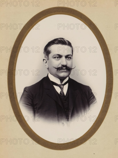 Portrait of a man, album cover, photograph, cabinet photograph, This photo is part of an album., inscription: 'Yu Chong, PEINTRE SUR IVOIRE ET AGRANDISSEMENT, DES PHOTOGRAPHES, 79 RUE DES PANIERS 79, HANOÏ', annotation: 'Lori..e', photographer: Yu Chong, Hanoi, 1880 - 1940, paper, pasteboard, daylight gelatin silver print, height 135 mm x width 96 mm, historical persons, A sepia-toned oval portrait of a man shown from the chest upward, framed by a double oval border; he has neatly combed dark hair, a prominent curled mustache, and a clean-shaven lower face, with light skin and a neutral expression, wearing a dark three-piece suit with a high white collar and a patterned tie, set against a plain light background.