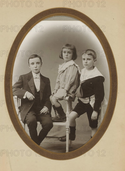 Group portrait of two boys and a young girl, album page, photograph, cabinet photograph, This photo is part of an album., inscription: 'A. Ligey, 21, Rue d'Auteuil, Paris', photographer: Albin Ligey, Paris, 1895 - 1920, Albin Ligey, photographer, paper, pasteboard, matte daylight collodion silver print, height 135 mm x width 96 mm, boy (child between toddler and youth), girl (child between toddler and youth), anonymous historical persons portrayed in a group, in a group-portrait, An oval framed sepia photograph showing three children posed on and around a wooden chair; on the left a boy in a dark suit and bow tie sits with one arm resting on the chair back and a neutral expression, in the center a girl with a bob haircut and a light dress with a ruffled collar sits on the chair seat with her hands resting on the rails and wearing dark boots, and on the right a boy in a dark sailor-style outfit with a light collar stands with one foot on the chair rung and a neutral expression.