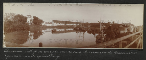 Panorama of the lookout and front of the old barracks in the Lower Town. Taken from the drawbridge., Panorama of the old barracks in the Lower Town of Batavia, seen from the drawbridge, album page, photograph, Part of Travel Album with photos of Batavia., Batavia, c. 1920, photographer, Java, West Java, paper, daylight gelatin silver print, height 248 mm x width 305 mm, barracks, prospect of city, town panorama, silhouette of city, overseas history, colonial history, Dutch East Indies, The, A wide sepia-toned riverside scene showing calm water reflecting buildings and boats, with a row of low whitewashed structures and a long gabled building along the far bank, a taller multi-story building with a tower and surrounding trees on the left, clustered small boats and masts moored near wooden sheds and piers on the right, a wooden railing and walkway in the foreground leading toward a small hut where a person is standing, and vegetation along the river edges under a pale sky.