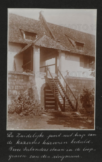 The southern gate with stairs of the barracks mentioned above. Two natives are standing in the trenches of the ring wall., Southern gate with stairs of the old barracks in Batavia, two Javanese are standing in the trenches of the ring wall, album page, photograph, Part of Travel Album with photos of Batavia., Batavia, c. 1920, photographer, Java, West Java, paper, daylight gelatin silver print, height 248 mm x width 305 mm, barracks, gate, entrance, overseas history, colonial history, Dutch East Indies, The, A sepia photograph of a two-story building with a tiled pitched roof and two small dormer openings, showing a covered porch supported by square columns and accessed by a wooden staircase with handrails leading down to an arched doorway set in a stone retaining wall; vegetation grows at the base of the wall and along the foreground, and a person is standing on the porch near the top of the stairs.