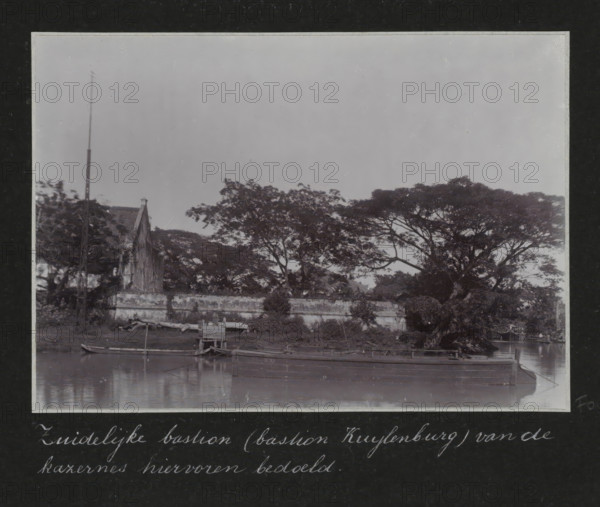 Southern bastion (Bastion Kuylenburg) of the barracks mentioned above., Southern Bastion Kuylenburg at the old barracks in Batavia, album page, photograph, Part of Travel Album with photos of Batavia., Batavia, c. 1920, photographer, Java, West Java, paper, daylight gelatin silver print, height 248 mm x width 305 mm, barracks, overseas history, colonial history, Dutch East Indies, The, A black and white photograph shows a waterfront scene with a calm body of water in the foreground and a low stone or concrete wall running parallel to the shore. Small wooden boats are moored at the water's edge, some tied to simple wooden structures and a short pier. Behind the wall, a cluster of large, spreading trees with dense canopies occupies much of the middle ground, their branches extending horizontally. To the left, a pitched-roof building with a visible gable and a tall, slender pole or mast stands partially obscured by foliage. The sky above is plain and light in tone.