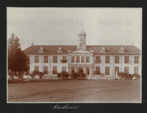Front of the Batavia City Hall, City Hall., album page, photograph, Part of Travel Album with Photos of Batavia., Batavia, c. 1920, photographer, Java, West Java, paper, daylight gelatin silver print, height 248 mm x width 305 mm, town hall, overseas history, colonial history, Dutch East Indies, The, A large three-story building with a central pedimented entrance and a columned portico extends horizontally across the image, flanked by symmetrical rows of shuttered windows and dormer windows on a tiled roof; a cupola with a domed roof and a slender spire rises from the center of the roofline above the entrance, and a row of low shrubs and taller palm-like plants lines the front of the building; trees frame the left and right edges, and a paved open area with visible rails or tracks runs in the foreground.