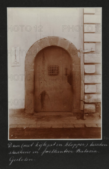 Closed door between the city hall and the post office in Batavia, on the left a sign of the ANIW, Door (with peephole and knocker) between city hall and post office Batavia. Closed., album page, photograph, Part of Travel album with photos of Batavia., Batavia, c. 1920, photographer, Java, West-Java, paper, daylight gelatin silver print, height 248 mm x width 305 mm, gate, entrance, overseas history, colonial history, Dutch East Indies, The, A weathered arched wooden door set into a stone surround within a pale wall, the door showing stains and a small barred opening near its upper center and a metal handle to one side, flanked on the right by a section of wall with deep rectangular block-like projections and on the left by a smooth plastered surface with a small rectangular plaque mounted on it, all seen above a worn stone threshold and paving.