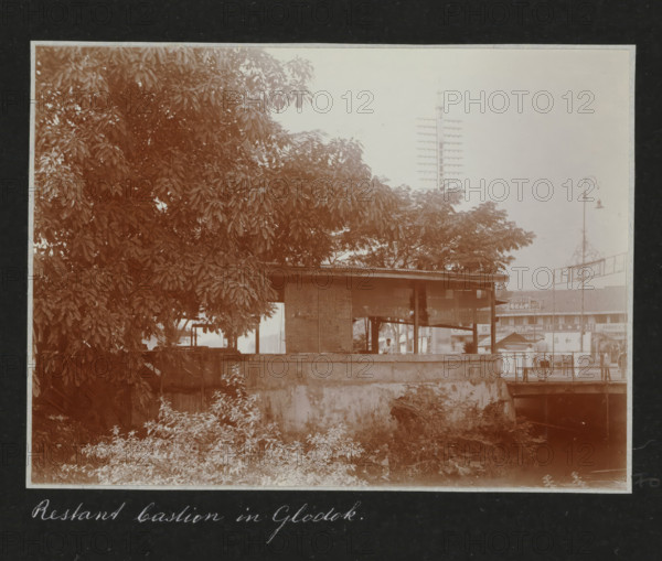 Remnant of a bastion in Glodok., Remnants of a bastion in the Chinese quarter of Glodok in Batavia, in the background modern traffic and advertising for a photo shop, album page, photograph, Part of a travel album with photos of Batavia., Batavia, c. 1920, photographer, Java, West Java, paper, daylight gelatin silver print, height 248 mm x width 305 mm, ruin of a dwelling, house, castle, studio, overseas history, colonial history, Dutch East Indies, The, A sepia-toned scene showing a large leafy tree occupying the left and center foreground with dense foliage, a low concrete wall in front of an open-sided wooden shelter with a corrugated roof and woven panels, a person partially visible sitting inside the shelter, vegetation and low plants growing along the base of the wall, a narrow waterway or dark channel at the right edge beneath a small bridge, a tall vertical pole with multiple horizontal crossarms rising behind the trees, a decorative street lamp on a slender post near the right...
