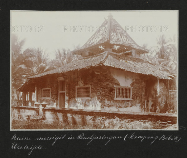 Remains of a mosque (west side) in Pendjaringang (kampong Bèbèk), Ruin of a mosque in Pendjaringang (kampong Bebek) West side., album page, photograph, Part of Travel album with photos of Batavia., Batavia, c. 1920, photographer, Java, West Java, paper, daylight gelatin silver print, height 248 mm x width 305 mm, ruin of a dwelling, house, castle, overseas history, colonial history, Dutch East Indies, The, A weathered brick building with a tiled, multi-tiered roof showing broken and missing tiles and exposed rafters, windows with vertical wooden bars, a narrow doorway, peeling plaster on the exterior walls, a low brick and stucco perimeter wall in front, and tall palm trees visible behind and beside the structure under a pale sky.