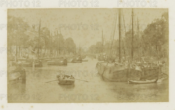 View of the Nieuwehaven in Rotterdam, photograph, Part of Photo album with cityscapes of Rotterdam., inscription: 'Nieuwehaven', Rotterdam, c. 1880 - c. 1900, photographer, paper, albumen print, height 108 mm x width 186 mm, rowing-boat, canoe, sailing-ship, sailing-boat, harbour (view, vehicle, ship, etc.), A sepia-toned scene of a narrow waterway lined with moored sailing vessels on both sides, showing tall masts and rigging, hulls with visible planks and ropes, and smaller rowboats on the water including one with a person rowing toward the foreground; tree-lined quays and dockside buildings are visible behind the boats, and the water surface reflects the vessels and ripples gently.